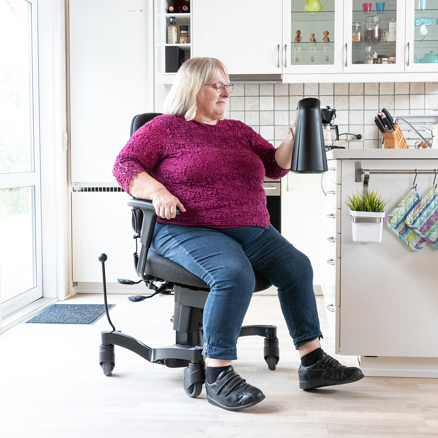 Woman using a mobility chair as a walking aid in her kitchen while carrying a coffee pot, showing how walking aids can support safe movement and independence in daily activities.